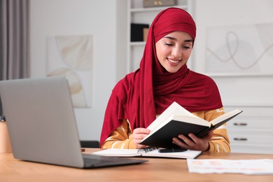 Muslim woman studying near laptop at wooden table in room