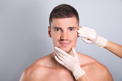 Doctor examining man's face for cosmetic surgery on grey background