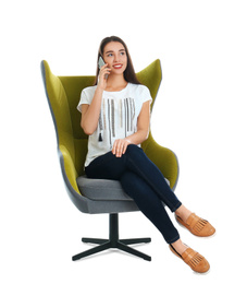 Young woman sitting in armchair and talking by smartphone on white background