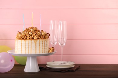 Photo of Caramel drip cake decorated with popcorn and pretzels near balloons and tableware on wooden table, space for text