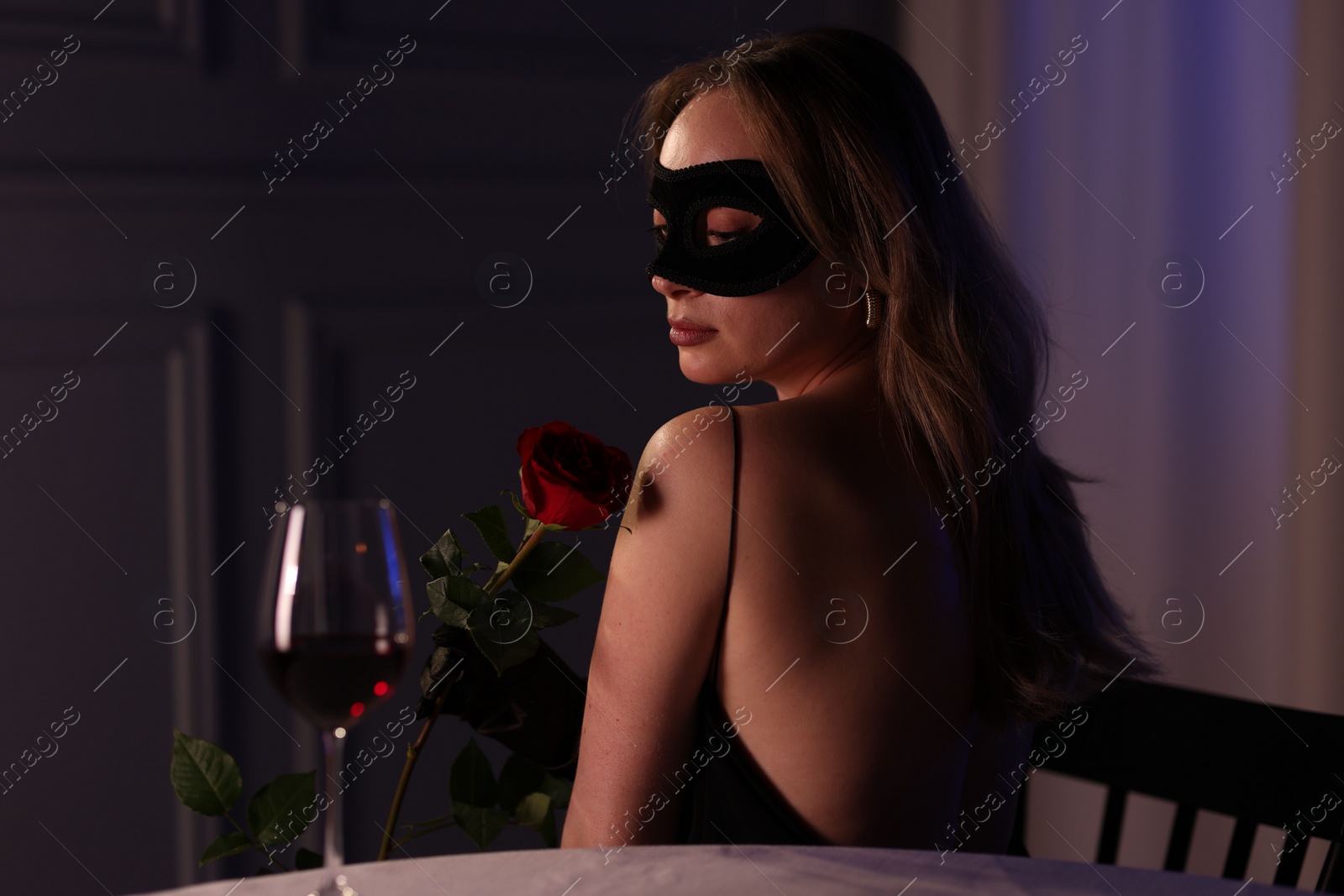 Photo of Elegant woman in black eye mask with rose indoors in evening