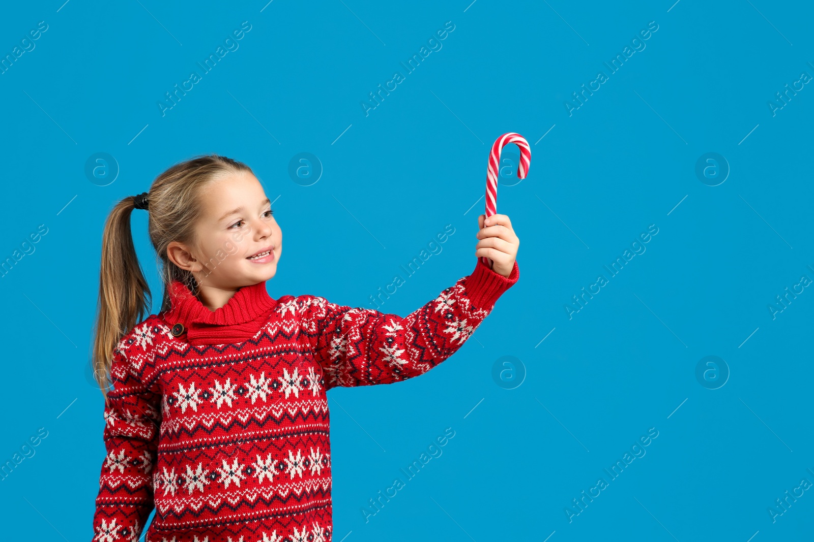 Photo of Cute little girl in knitted Christmas sweater holding candy cane on blue background, space for text
