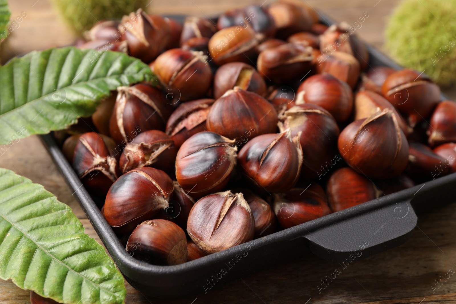 Photo of Delicious roasted edible chestnuts and green leaves on wooden table, closeup