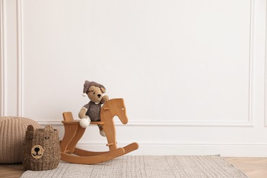 Photo of Rocking horse with bear toy, pouf and wicker basket near white wall in child room, space for text. Interior design