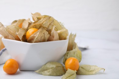 Ripe physalis fruits with calyxes in bowl on white marble table, closeup. Space for text
