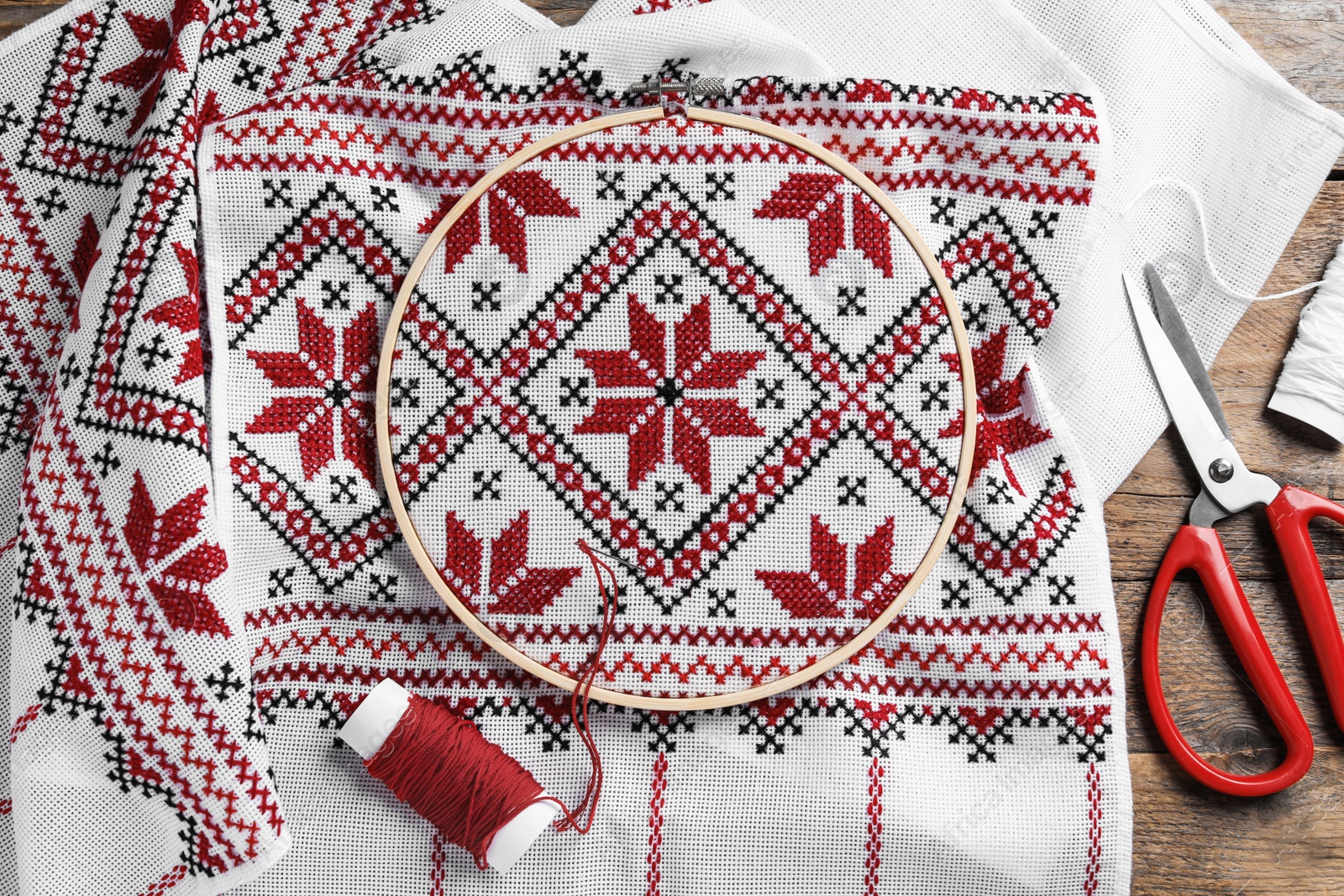 Photo of White fabric with red Ukrainian national embroidery in hoop, needle, scissors and thread on wooden table, flat lay