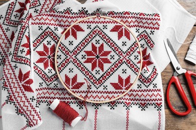 White fabric with red Ukrainian national embroidery in hoop, needle, scissors and thread on wooden table, flat lay