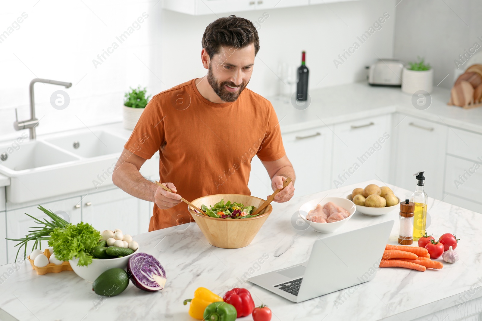 Photo of Man making dinner while watching online cooking course via laptop in kitchen