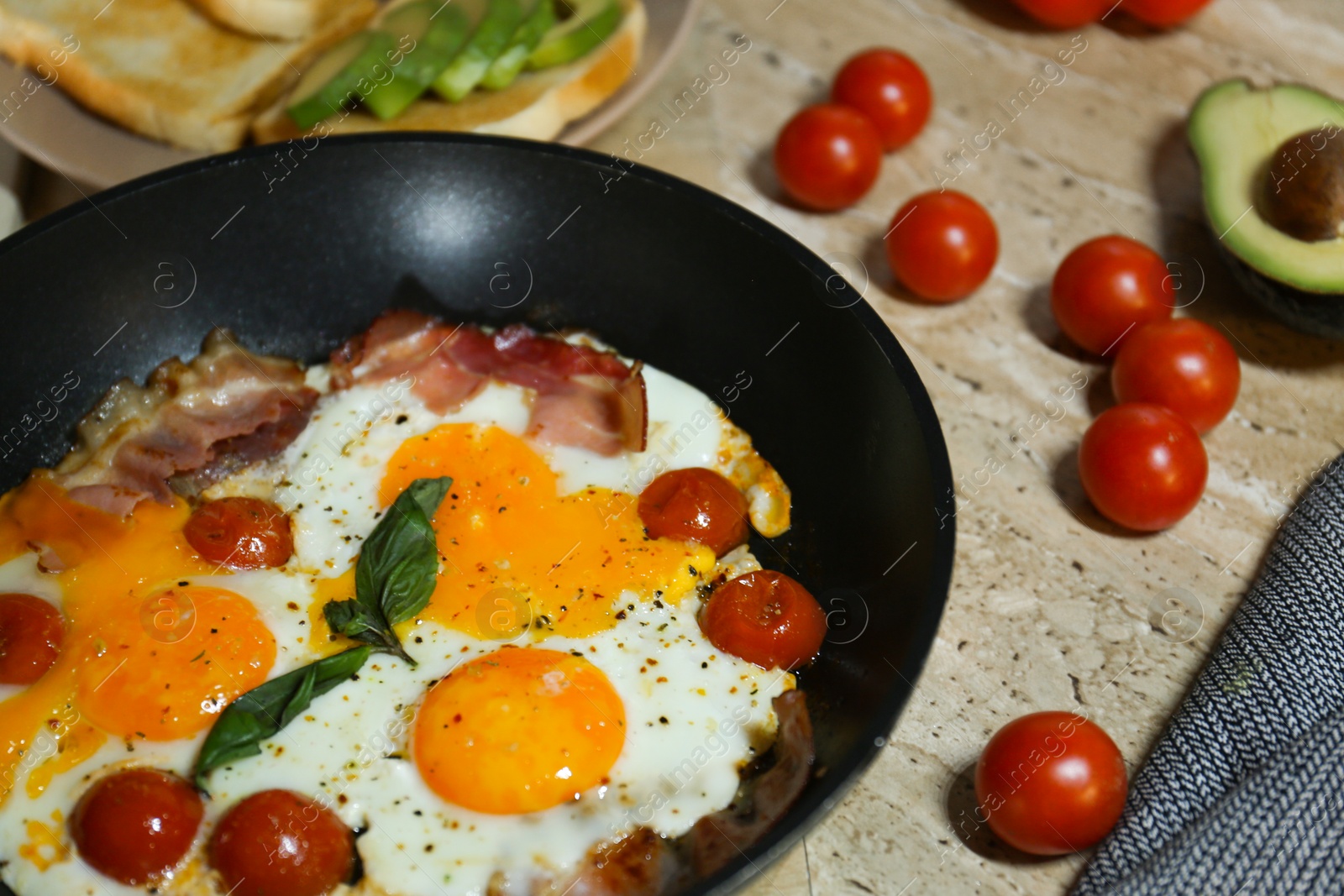 Photo of Fried with eggs, bacon and tomatoes in frying pan near ingredients on wooden table, closeup. Tasty breakfast