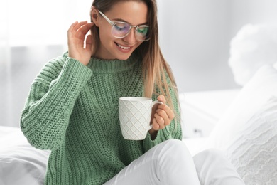 Photo of Young woman in warm sweater with hot drink on bed at home