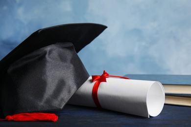 Graduation hat, books and student's diploma on dark blue wooden table against light blue background