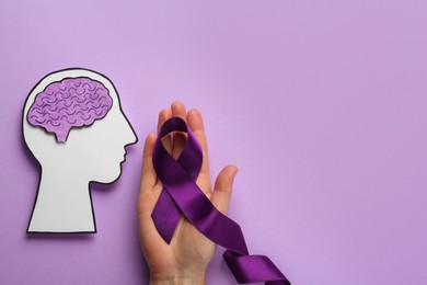Woman holding purple ribbon near paper human head cutout over violet background, top view. Epilepsy awareness