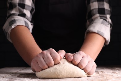 Man kneading dough at wooden table on dark background, closeup