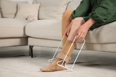 Photo of Woman putting on compression tights with stocking donner in living room, closeup. Prevention of varicose veins