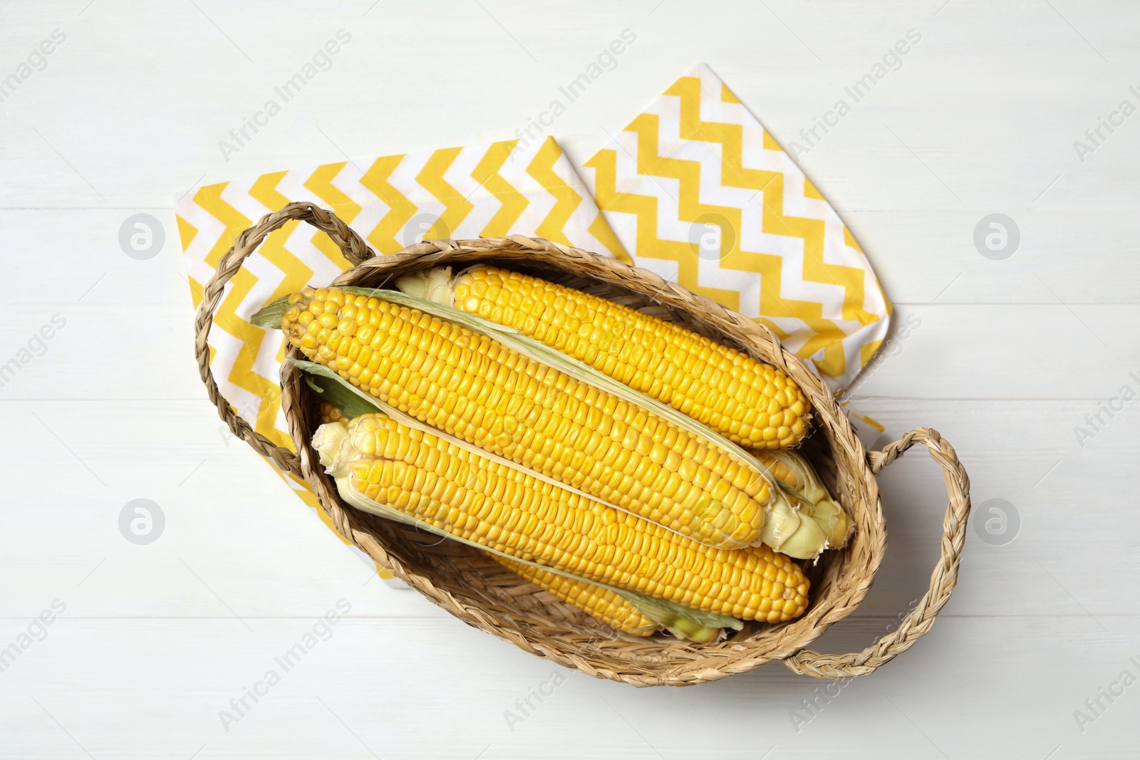 Photo of Basket of corn cobs on white wooden table, flat lay