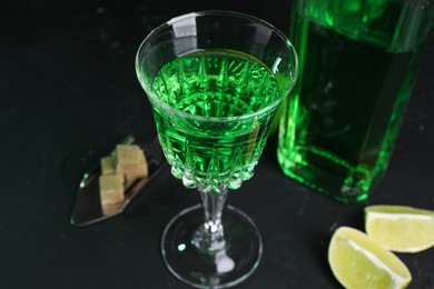Photo of Absinthe in glass, spoon, brown sugar and lime on black table, closeup. Alcoholic drink