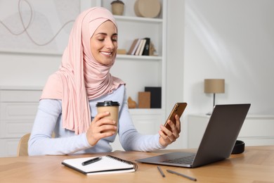 Photo of Muslim woman with cup of coffee using smartphone near laptop at wooden table in room