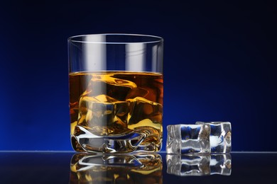 Photo of Whiskey in glass with ice cubes on table against dark blue background, closeup