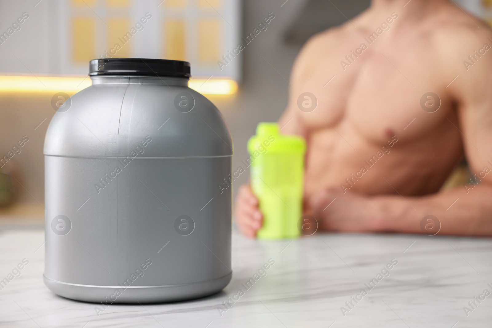 Photo of Young man with shaker of protein at white marble table in kitchen, focus on jar of powder