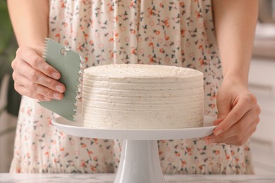 Photo of Woman using scraper to decorate cake at white table, closeup