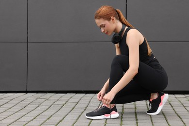 Beautiful woman in stylish gym clothes with headphones tying shoelace of sneakers near dark grey wall on street, space for text