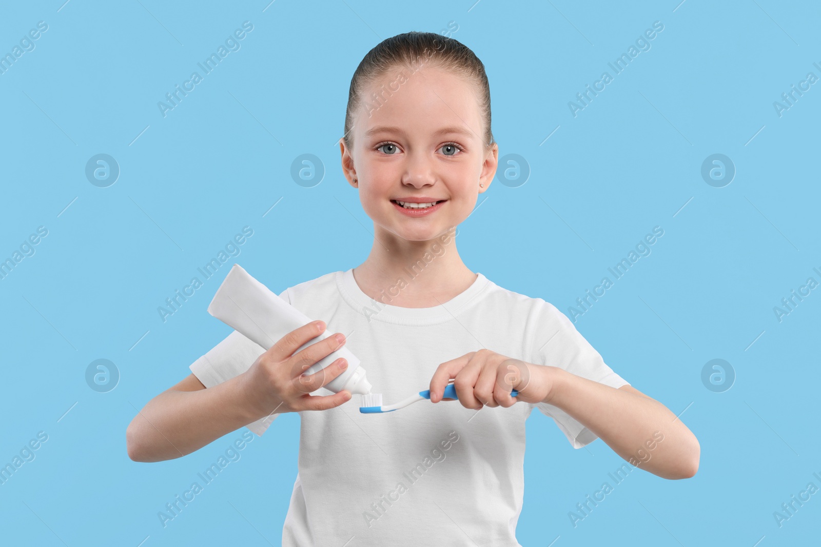 Photo of Happy girl squeezing toothpaste from tube onto toothbrush on light blue background