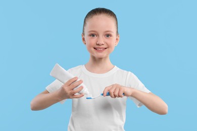 Happy girl squeezing toothpaste from tube onto toothbrush on light blue background