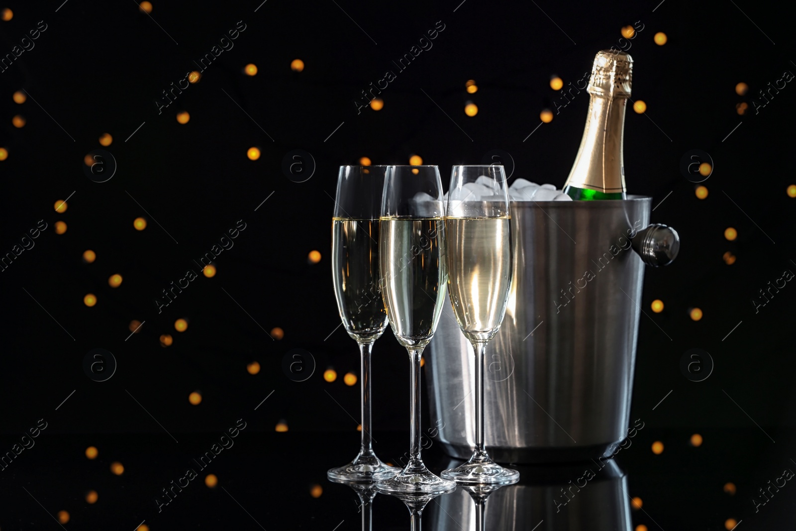 Photo of Glasses with champagne and bottle in bucket on dark table against blurred lights