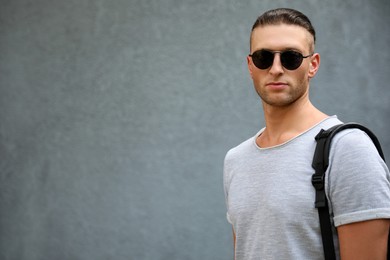 Photo of Handsome young man in stylish sunglasses with backpack near grey wall, space for text