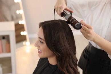 Photo of Hairdresser working with beautiful woman using curling hair iron in salon