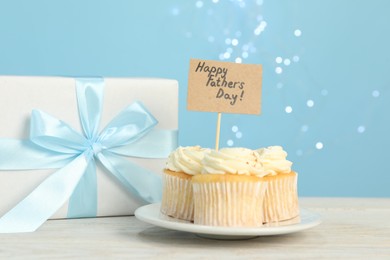 Photo of Happy Father's Day. Tasty cupcakes with greetings and gift box on light wooden table, closeup