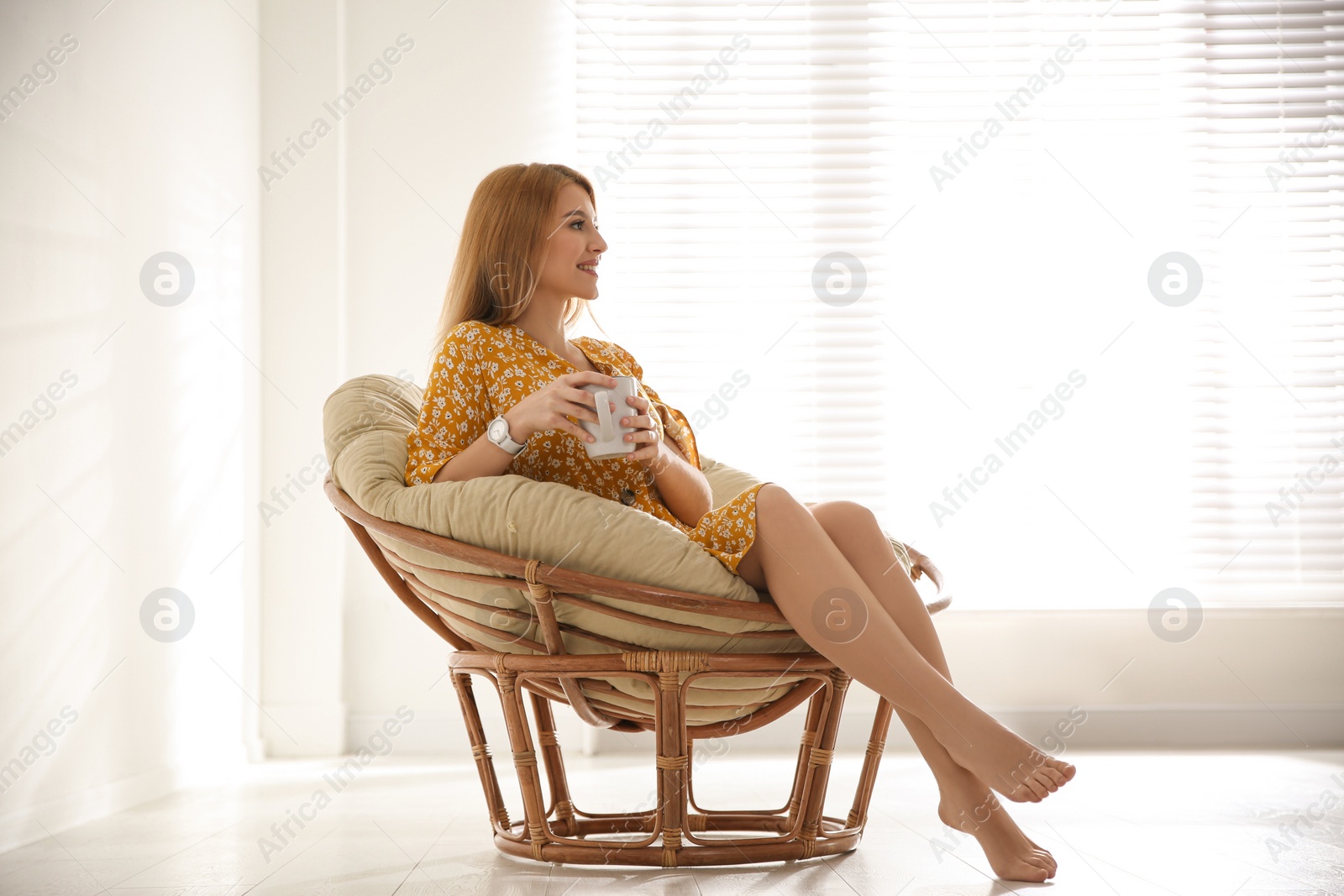 Photo of Young woman with cup of drink relaxing in papasan chair near window at home