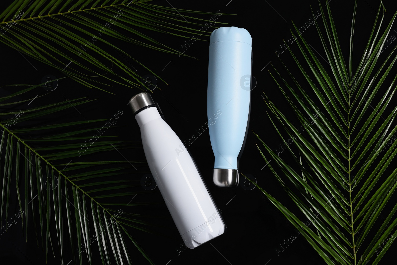 Photo of Thermo bottles and green leaves on black background, flat lay