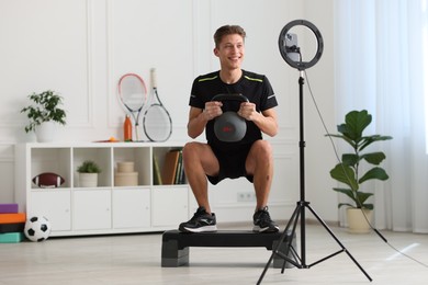 Photo of Smiling sports blogger training with kettlebell while streaming online fitness lesson at home