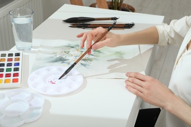 Woman painting flowers with watercolor at white table indoors, closeup. Creative artwork