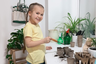 Photo of Little girl adding soil into peat pots on window sill indoors. Growing vegetable seeds