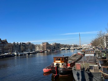 Amsterdam, Netherlands - March 01, 2023: Picturesque view of river embankment with moored boats in city under blue sky