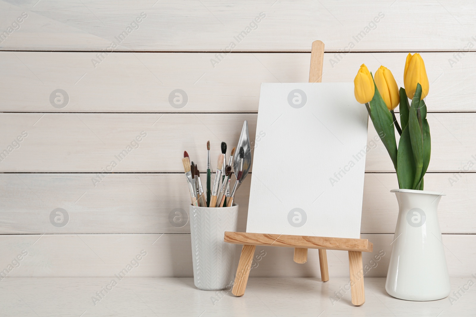 Photo of Easel with blank canvas, brushes and tulips on white wooden table. Space for text