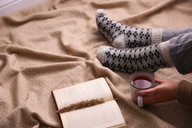 Photo of Woman with cup of hot winter drink and book on warm plaid, closeup. Space for text