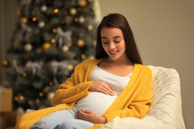 Happy pregnant woman in armchair at room decorated for Christmas. Expecting baby