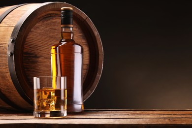 Whiskey with ice cubes in glass, bottle and barrel on wooden table against black background, space for text