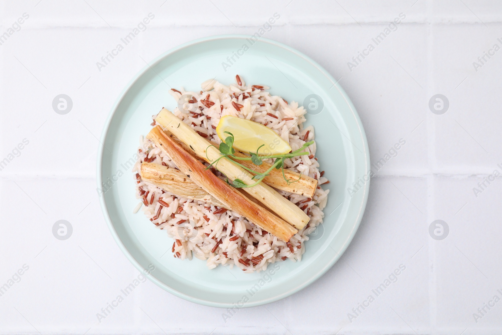 Photo of Plate with baked salsify roots, lemon and rice on white tiled table, top view