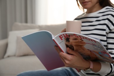 Photo of Woman with cup of drink reading sports magazine at home, closeup