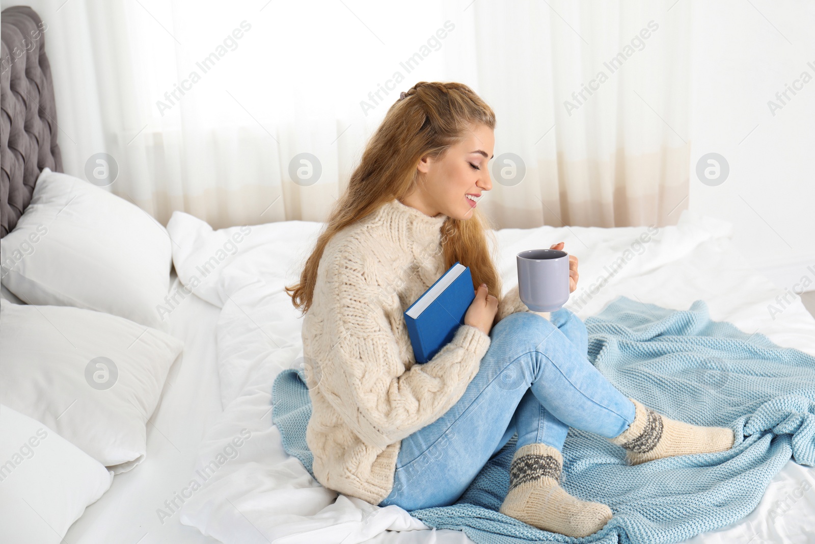 Photo of Attractive young woman in cozy warm sweater with cup of hot drink and book sitting on bed at home