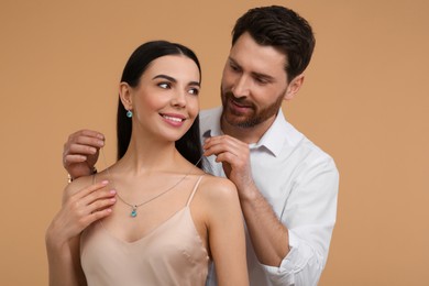 Man putting elegant necklace on beautiful woman against beige background