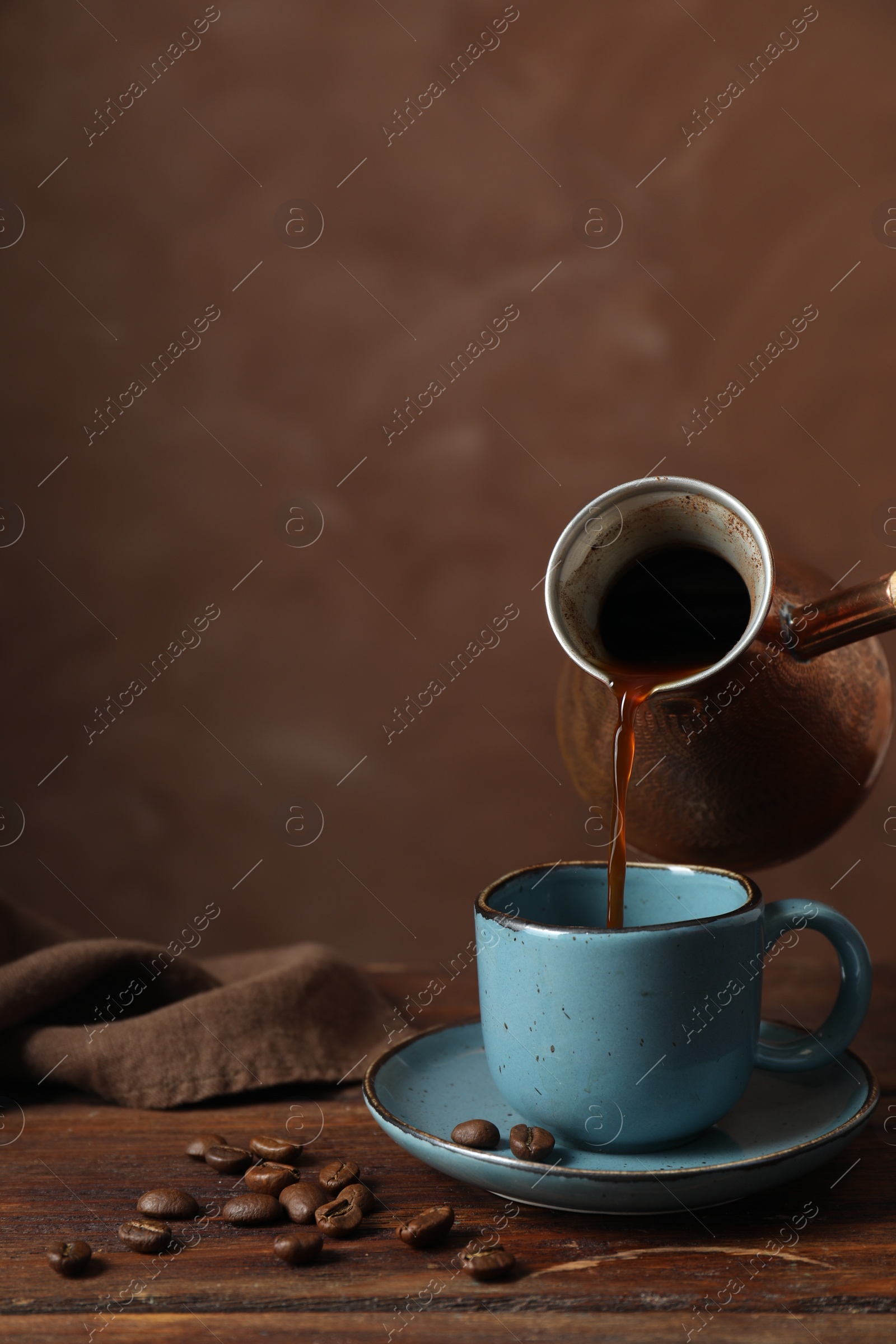 Photo of Turkish coffee. Pouring brewed beverage from cezve into cup at wooden table against brown background, space for text