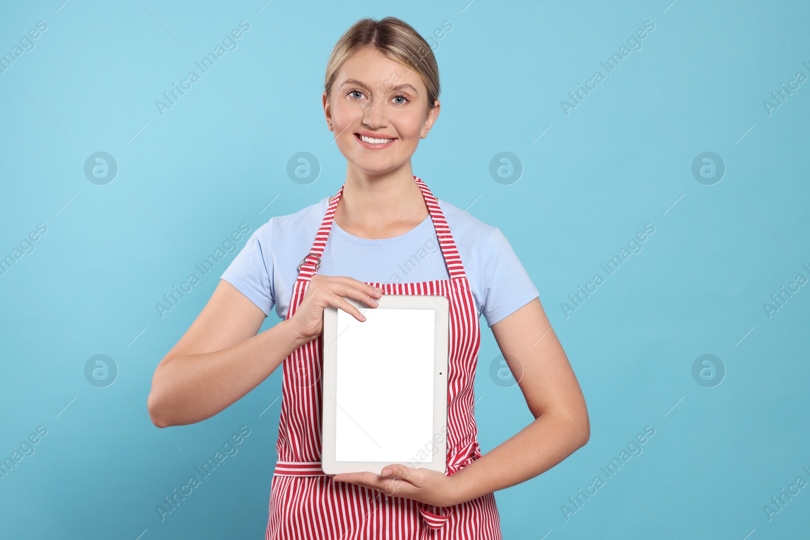 Photo of Beautiful young woman in clean striped apron with tablet on light blue background