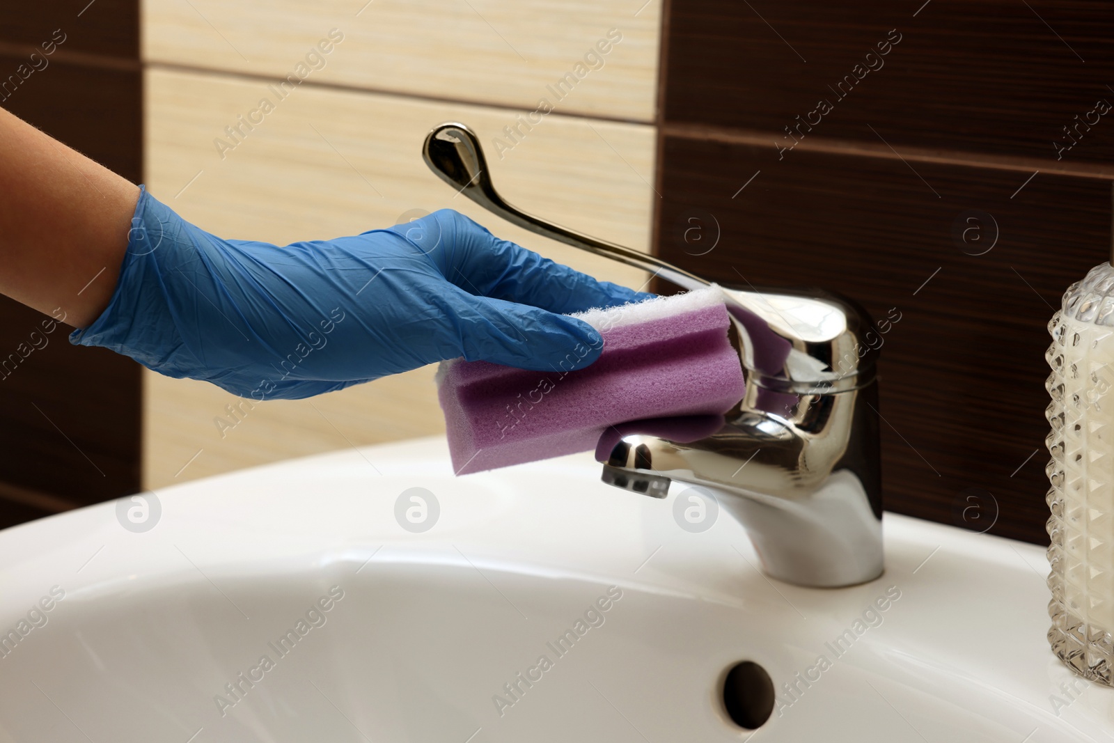 Photo of Woman in gloves cleaning faucet of bathroom sink with sponge, closeup