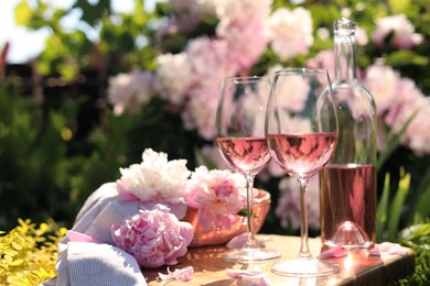 Bottle and glasses of rose wine near beautiful peonies on wooden table in garden