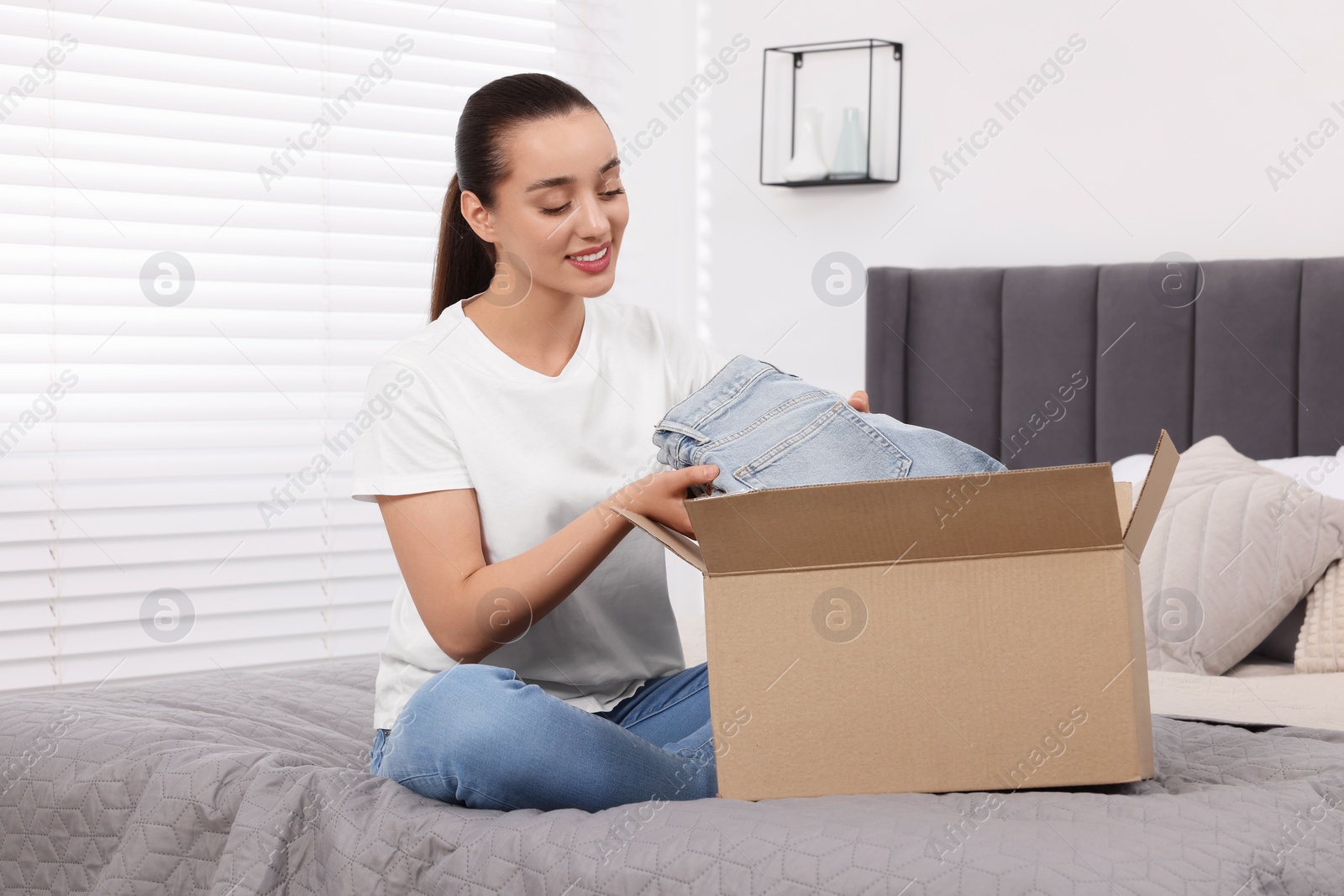 Photo of Happy young woman taking jeans out of box on bed at home. Online shopping